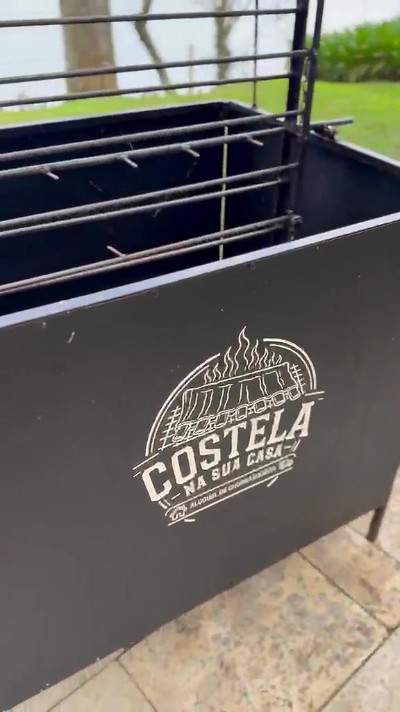Churrasqueira Rústica preta com o logo da Costela na Sua Casa na lateral, em um terraço à beira de um lago